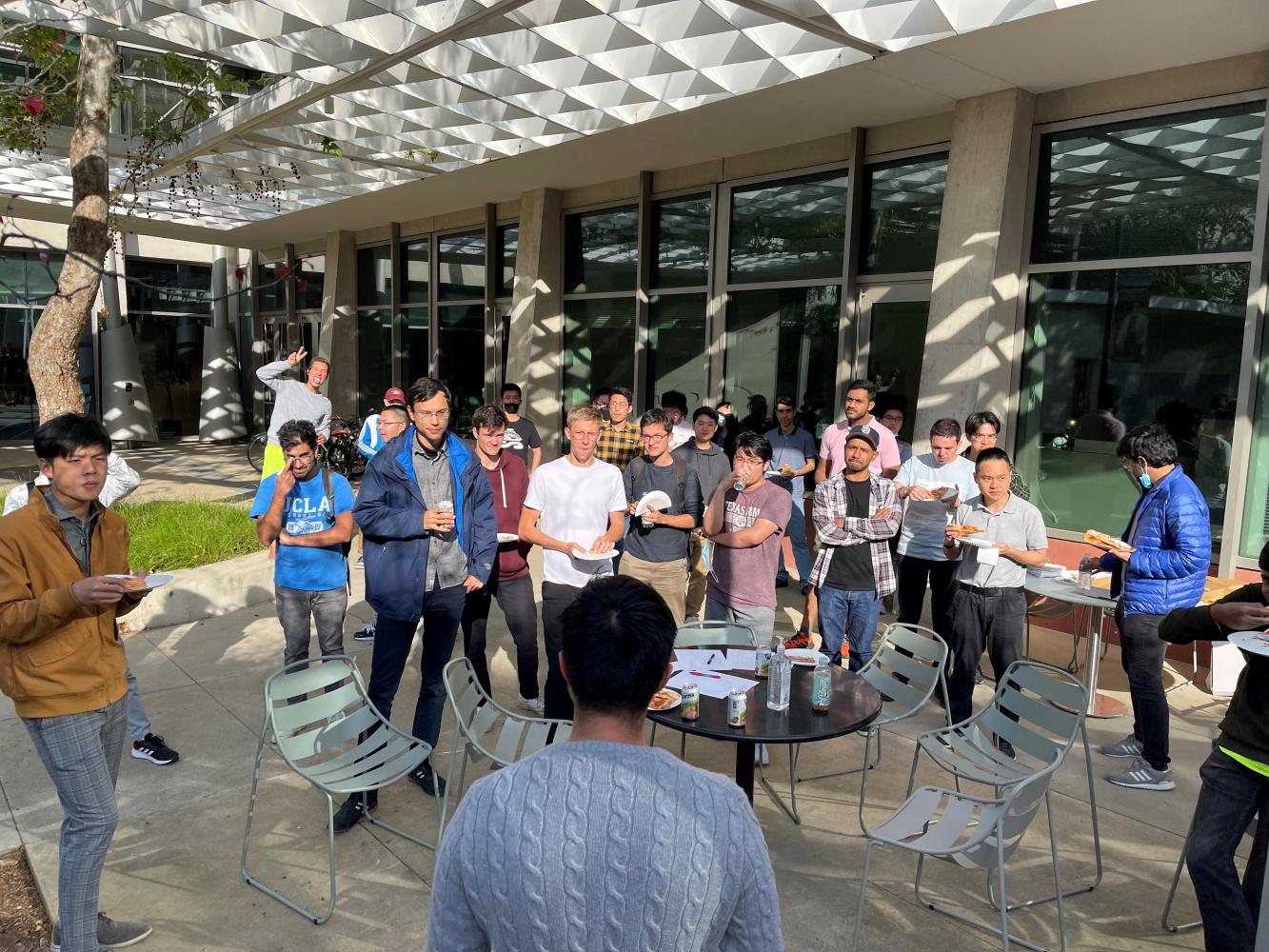 Picture of a group of about 25 RoboGrads members standing and eating pizza while someone give an announcement