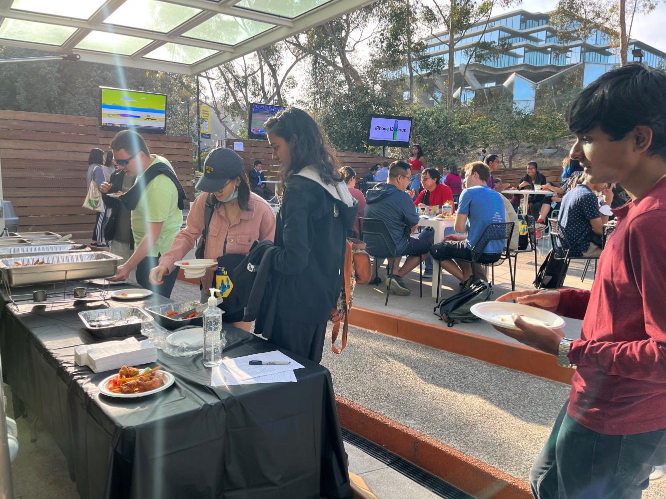 Picture of the RoboGrads end of the year social with people serving themselves food. There are also groups of people eating and talking at tables, and Geisel Library is in the background.